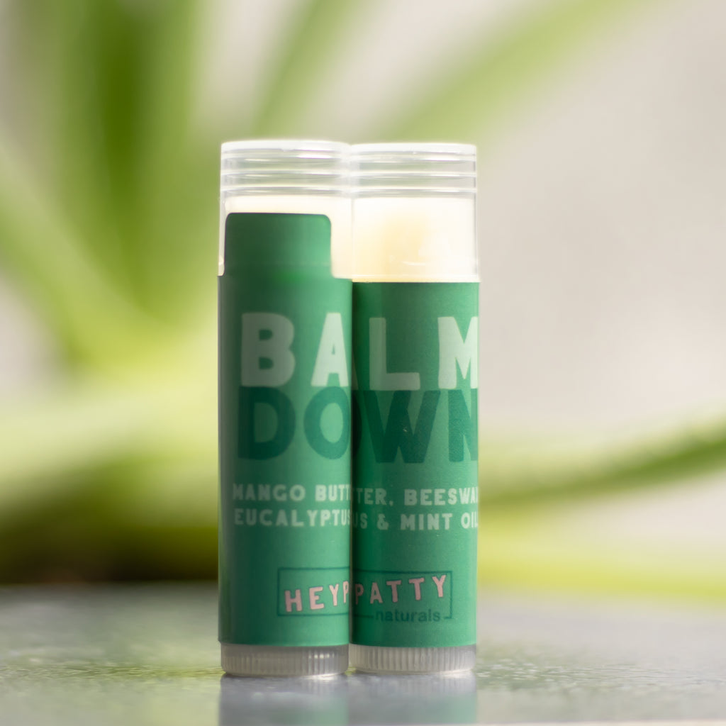 Two green balm sticks with HEYPATTY Naturals branding on a blurred green background