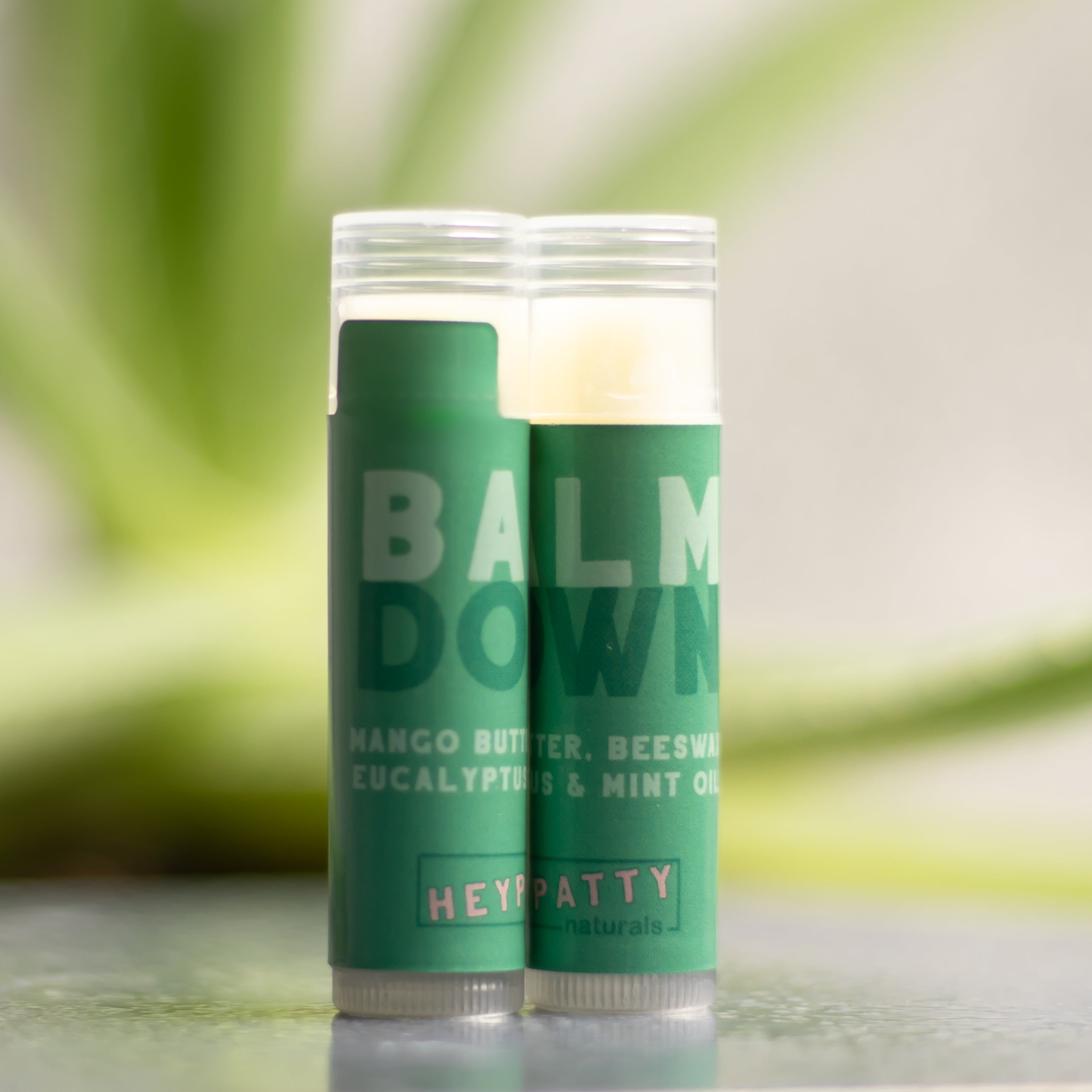 Two green balm sticks with HEYPATTY Naturals branding on a blurred green background