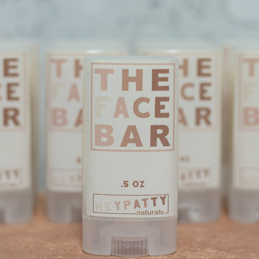 Small containers labeled 'The Face Bar' by HEYPATTY Naturals on a stone surface.